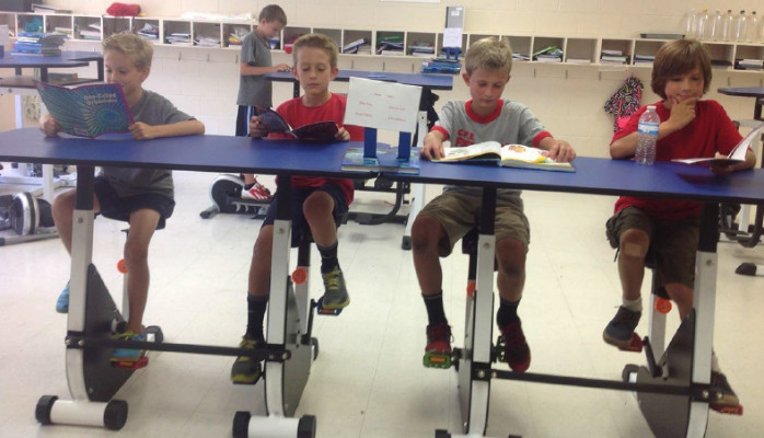 Photo of four young boys sitting at a table with peddles underneath. This photo is associated with an article on the 1st fully kinesthetic classroom in the USA.