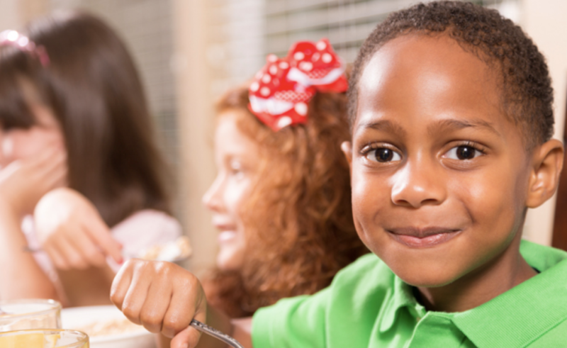 Photo of a young boy in a green shirt eating in a school cafeteria. There are two young girls in the background. This photo is associated with the article about Washington DC fully funding school breakfasts.